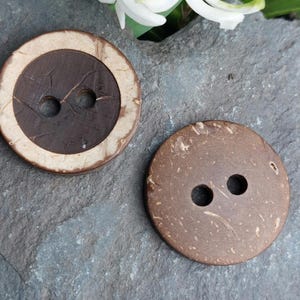May include: Two large, round coconut buttons. One button has a dark brown centre with a light brown outer ring. The other button is a solid, medium brown colour. Both buttons have two holes for sewing.