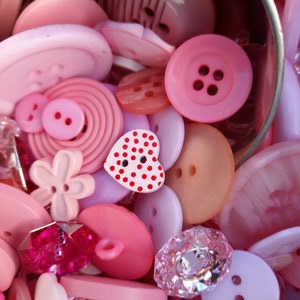 May include: A collection of pink, white and red buttons in various shapes and sizes. The buttons are in a metal container. Some buttons have a polka dot pattern.