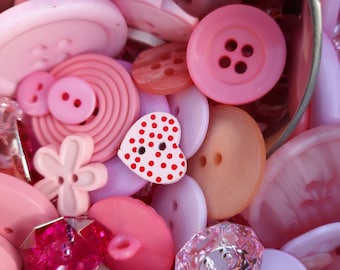 May include: A collection of pink, white, and red buttons in various shapes and sizes. The buttons are in a metal container. Some buttons have a polka dot pattern.