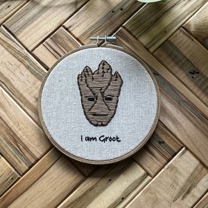 May include: Embroidered hoop art featuring a brown and tan Groot character from Guardians of the Galaxy. The hoop is white with the text "I am Groot" in black.