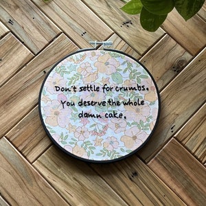 May include: Round embroidery hoop with floral fabric and the text "Don't settle for crumbs. You deserve the whole damn cake." The fabric has a light pink background with peach and yellow flowers. The hoop is on a wooden surface.