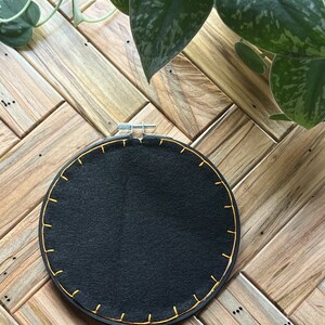 May include: A black embroidery hoop with a yellow stitched border. The hoop is on a wooden surface.