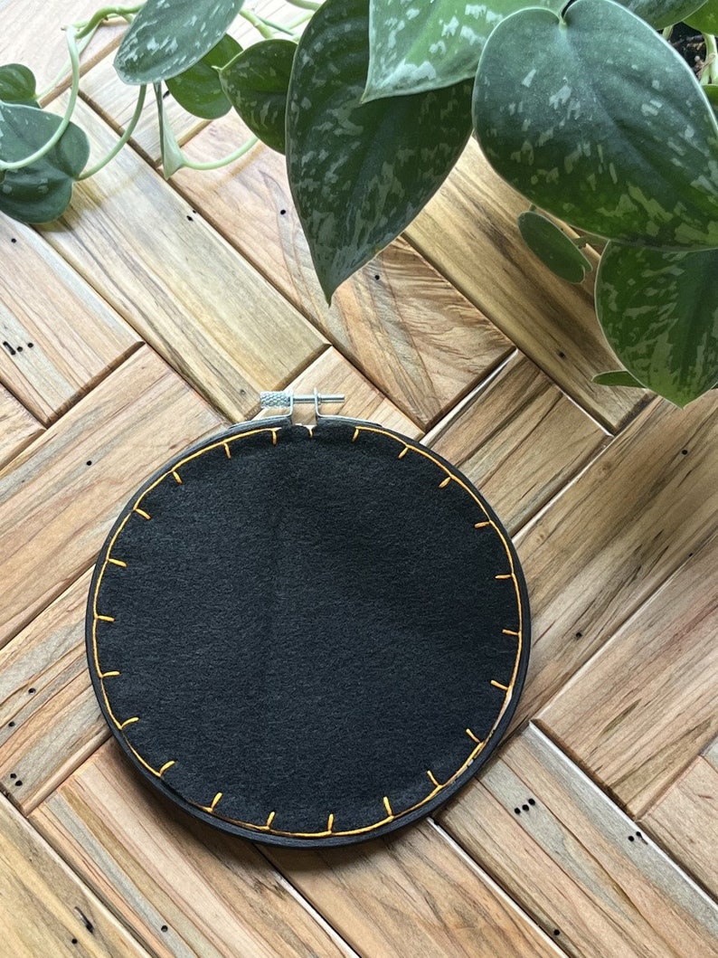 May include: A black embroidery hoop with a yellow stitched border. The hoop is on a wooden surface.
