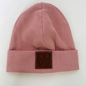 Könnte beinhalten: Ein altrosa Beanie-Mütze mit Rippstruktur. Ein braunes quadratisches Patch mit dem Wort "LOVE" ist auf der Vorderseite aufgenäht. Die Mütze ist aus weichem Strickmaterial gefertigt.