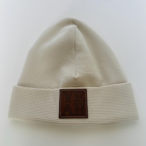 Könnte beinhalten: Eine beige Beanie-Mütze aus Waffelstrick mit einem braunen Lederaufnäher. Der Aufnäher zeigt das Wort "YEAH" in Großbuchstaben. Die Mütze hat einen umgeschlagenen Saum und eine strukturierte Oberfläche. Die Mütze ist neutralfarben.
