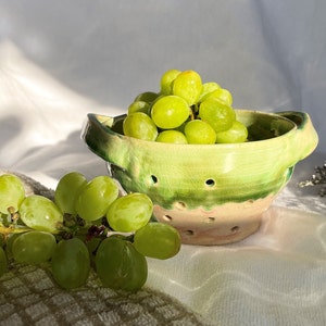 May include: A green and white ceramic colander with a handle on each side, filled with green grapes. The colander is sitting on a white surface with a brown towel underneath.