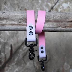 Puede incluir: Dos correas rosas con cierres giratorios de metal negro y detalles en lila claro. Las correas están unidas con remaches plateados. El fondo es una viga de madera envejecida y una superficie gris texturizada.