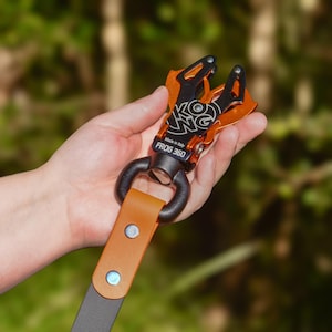 May include: A black and orange metal Frog 360 carabiner with a leather strap. The carabiner has the text "KONG" and "Made in Italy" printed on it. The strap is brown with two silver rivets.