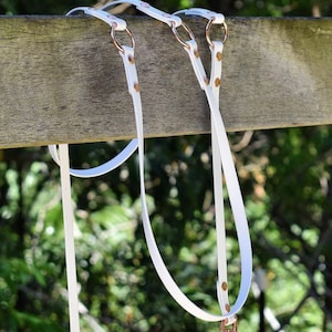 Puede incluir: Correa de perro de cuero blanco con herrajes de cobre y dos anillos. La correa está colgada de una valla de madera.