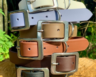 May include: A stack of five dog collars in different colors, including white, lavender, brown, orange, and black. Each collar has a silver buckle and a silver ring.