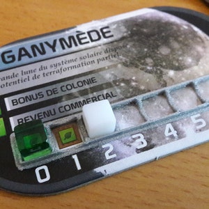 Może przedstawiać: Karta do gry z tekstem "GANYMÈDE" i "La plus grande lune du système solaire dispose d'un potentiel de terraformation partiel". Karta ma białą płytkę na torze numerowanym od 0 do 6. Karta ma również dwie zielone ikony liści i zieloną płytkę.