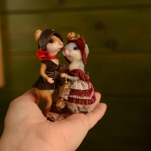 May include: Two small felted mouse figurines dressed in detailed clothing. One mouse wears a dark brown vest, hat, and red scarf. The other mouse wears a maroon bonnet, a striped skirt with lace trim, and carries a small gold-toned lantern accessory.
