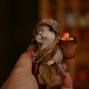 May include: A small, anthropomorphic mouse figurine wearing a textured brown hat and a tan tunic. The mouse carries a brown woven basket on its back filled with small orange fruits and has a small gray fish-shaped accessory hanging at its side.