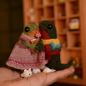 Op de afbeelding: Twee gebreide kikker knuffels die een klein boek en een bloem vasthouden. De kikker links draagt een roze en paarse jurk. De kikker rechts draagt een groene, rode en blauwe trui. Beide kikkers houden kleine witte keramische beeldjes vast.