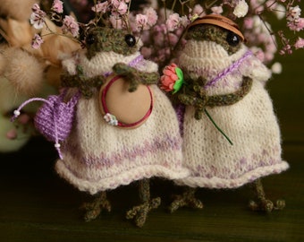 Puede incluir: Dos muñecas de rana de crochet que llevan vestidos y sombreros. Una rana sostiene una pequeña flor rosa y la otra sostiene un sombrero marrón. Las ranas están de pie sobre una superficie marrón oscura.