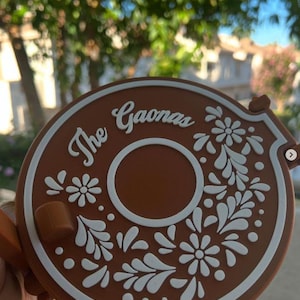 May include: A brown circular object with the text "The Gaonas" and white floral designs. The object has a handle and a central hole. The background is blurred with green foliage.
