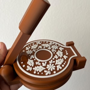 May include: A brown castanet with a handle. The castanet has a circular design with white floral patterns and a white border. The handle is rectangular and angled. The castanet is held in a hand.