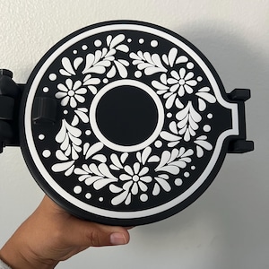 May include: Black and white decorative round object with a floral design. The object has a central black circle and a white floral pattern around the edge. The object is held by a hand.