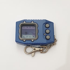 May include: A blue and silver digital pet device with a screen displaying a pixelated image. The device has three buttons and a silver chain attached.