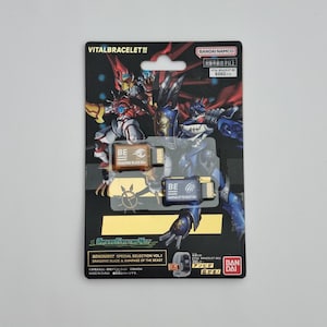 Może przedstawiać: Zestaw akcesoriów Vital Bracelet BE z dwiema kartami pamięci BE. Opakowanie jest czarne z kolorowymi ilustracjami postaci. Karty są oznaczone jako "Dragonic Blaze" i "Rampage of the Beast". Widoczne jest logo Bandai Namco.