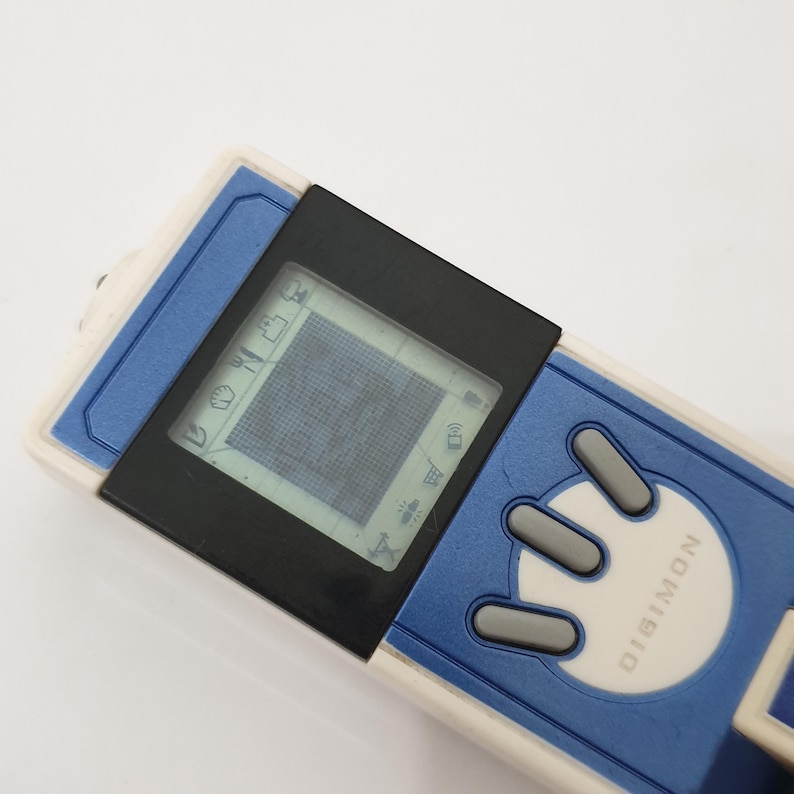 May include: A blue, white, and black Digimon digital pet toy. The device has a small screen displaying pixelated graphics and several gray buttons. The word "DIGIMON" is printed on the device.