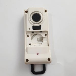 May include: A white electronic device with a black circular lens and a black U-shaped handle. The device has a rectangular shape with several screws and a small red light. The top section has a circular black lens.