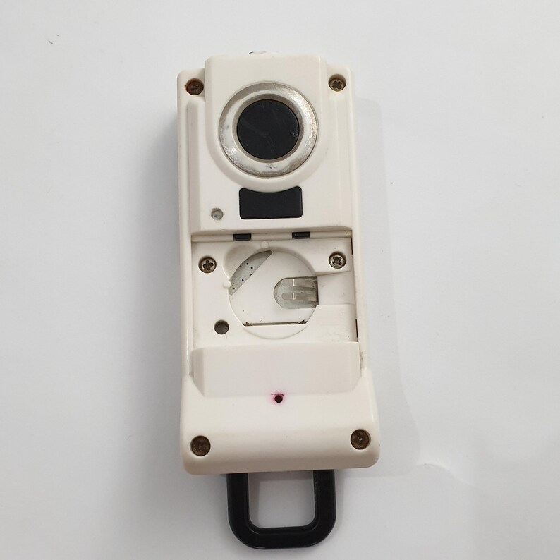 May include: A white electronic device with a black circular lens and a black U-shaped handle. The device has a rectangular shape with several screws and a small red light. The top section has a circular black lens.