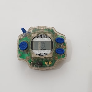May include: A clear plastic toy with a green circuit board, a digital display, and two blue buttons. The display shows the text "JUMPIN'" and a number.
