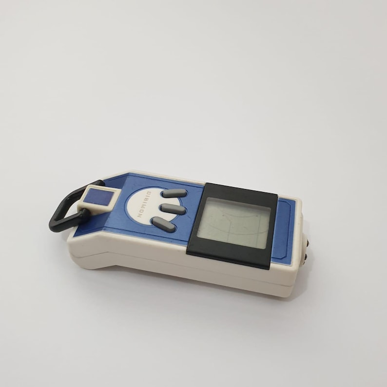 May include: A blue, white, and black Digimon digital pet toy. The handheld device features a small screen, three gray buttons, and the word "Digimon" printed on the front. A black loop is attached to the top.
