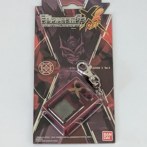 May include: A red and black Digimon X Ver.2 digital pet toy on its original packaging. The packaging features Japanese text and a graphic of a Digimon character. The toy has a silver keychain attached.