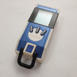 May include: A blue and white Digimon digital pet toy. The toy has a rectangular shape with a screen, three gray buttons, and the word "DIGIMON" printed on the front. A black loop is attached to the bottom.