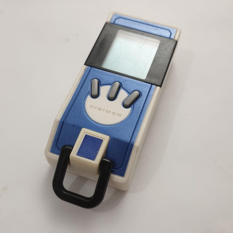 May include: A blue and white Digimon digital pet toy. The toy has a rectangular shape with a screen, three gray buttons, and the word "DIGIMON" printed on the front. A black loop is attached to the bottom.