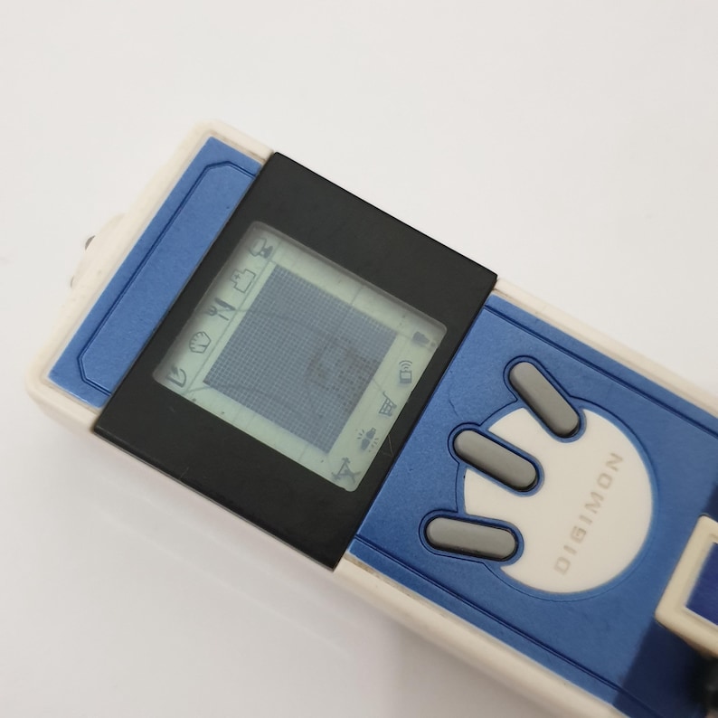 May include: A blue and white Digimon digital pet toy with a black screen displaying icons. The device has three gray buttons and the word "DIGIMON" printed on the front. The toy is rectangular and has a small screen.
