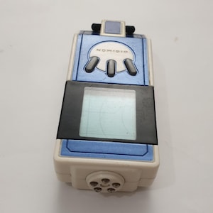 May include: A vintage Digimon digital pet device in white, blue, and black. The device has a rectangular screen, a series of buttons, and the word "DIGIMON" printed on the top. The device is a collectible item.