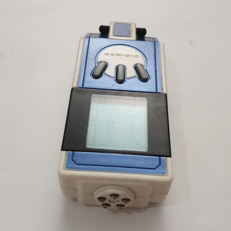 May include: A vintage Digimon digital pet device in white, blue, and black. The device has a rectangular screen, a series of buttons, and the word "DIGIMON" printed on the top. The device is a collectible item.
