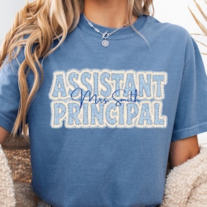May include: A blue short-sleeve t-shirt featuring the text "ASSISTANT PRINCIPAL" in a light blue and white patterned font, with the script text "Mrs Smith" centered in dark blue. The shirt has a crew neckline and a relaxed fit.