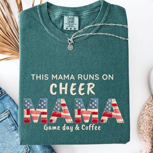 May include: Blue Spruce colored sweatshirt with the text "THIS MAMA RUNS ON CHEER MAMA Game day & Coffee". The word "MAMA" is designed with a red, white, and blue American flag pattern. A silver necklace with a pendant is on the shirt.