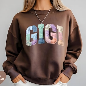 May include: Dark chocolate brown sweatshirt with the word "GIGI" in patchwork letters across the chest. The letters are made of various patterned fabrics in shades of blue, green, purple, and pink. The sweatshirt has a crew neck and long sleeves.