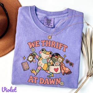 May include: A violet short-sleeve t-shirt with a graphic print of a patchwork frog carrying a tote bag, a clock, and books. The text reads WE THRIFT AT DAWN. The shirt features a crew neckline and a Comfort Colors label on the inside collar.