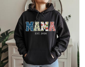 May include: Black hooded sweatshirt with the word "MAMA" in a decorative font. The letters are filled with festive patterns, including snowflakes, greenery, and stripes. The text "EST. 2026" is below the word "MAMA". The sleeve has the name "Rosie" embroidered.
