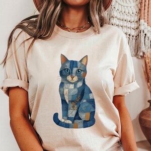 May include: A light beige short-sleeved t-shirt featuring a graphic of a sitting cat composed of various blue denim-style patchwork squares. The cat has bright blue eyes and a small golden sprig illustration on its chest area.