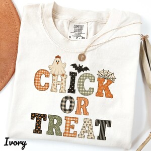 May include: An ivory t-shirt featuring the text CHICK OR TREAT in patterned block letters. The design includes a ghost-like chick, a black bat, and a spider web. The shirt has a Comfort Colors label at the neck and a gold-toned necklace rests on top.