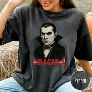 May include: Dark gray t-shirt featuring a portrait of Dracula with a black cape and bow tie. The word "DRACULA" is printed in red, dripping letters. The shirt is a relaxed fit.
