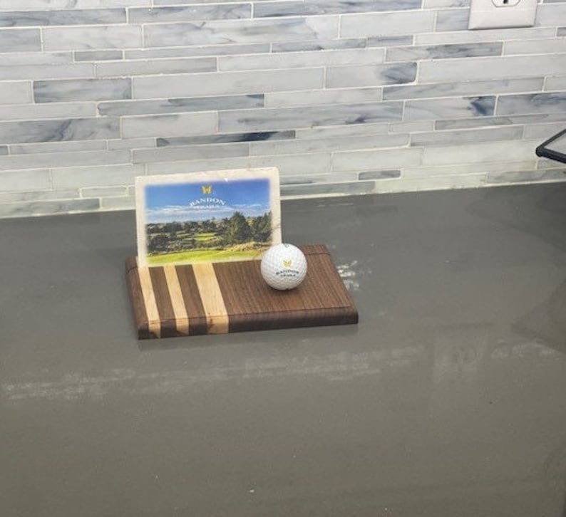 Pu&ograve; includere: Un tagliere di legno con un motivo a strisce in marrone e bianco. Una palla da golf e una cartolina con il testo "Wandoon Golf Club" sono sopra il tagliere.