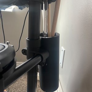 Op de afbeelding: Zwarte drumstickhouder bevestigd aan een zwarte drumstandaard. De houder is gemaakt van plastic en heeft een cilindrische vorm. De houder is met een metalen klem aan de standaard bevestigd.