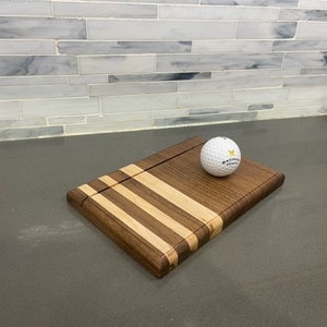 Pu&ograve; includere: Un tagliere in legno con un design in noce e acero. Il tagliere ha una scanalatura per raccogliere i succhi e una piccola palla da golf poggia sul tagliere.