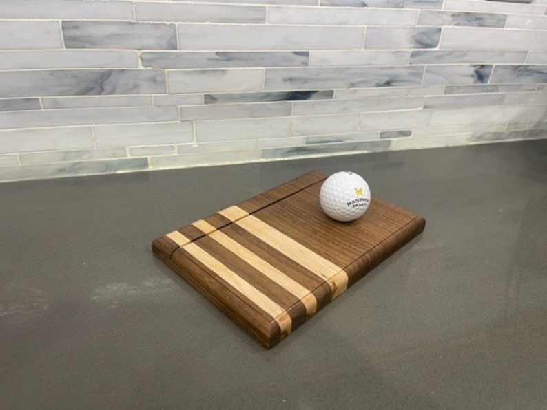 Pu&ograve; includere: Un tagliere in legno con un design in noce e acero. Il tagliere ha una scanalatura per raccogliere i succhi e una piccola palla da golf poggia sul tagliere.
