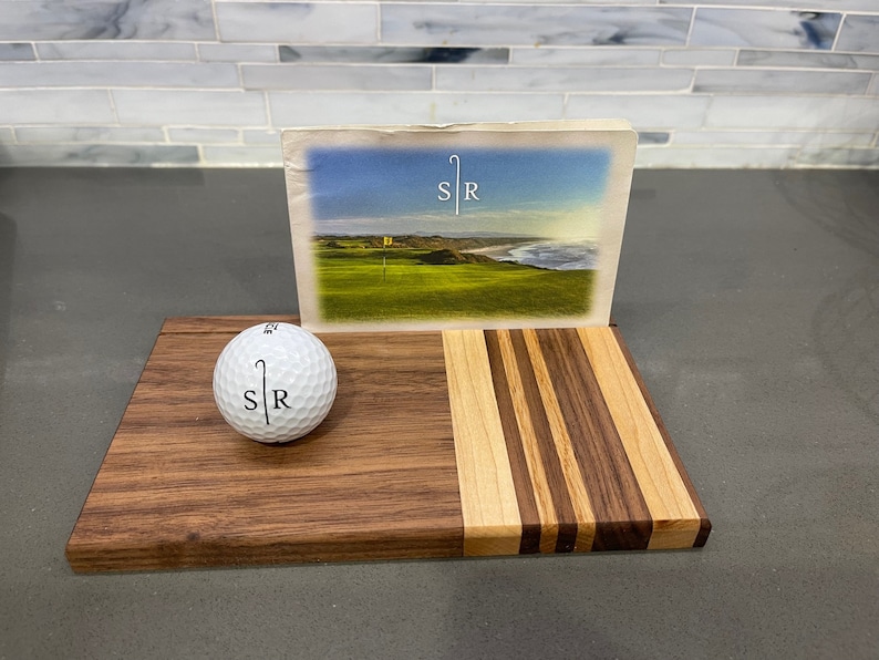 Pu&ograve; includere: Un tagliere di legno con un design a strisce. Il tagliere ha una palla da golf con le lettere "SR" su di esso e una cartolina con un campo da golf e le lettere "SR" su di esso.