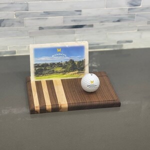 Pu&ograve; includere: Un tagliere di legno con un design a strisce in legno marrone e chiaro. Una palla da golf con il logo Bandon Dunes &egrave; sul tagliere. Un biglietto postale con il logo Bandon Dunes &egrave; dietro la palla da golf.