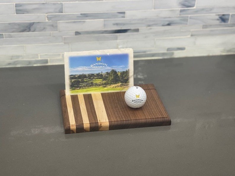 Pu&ograve; includere: Un tagliere di legno con un design a strisce in legno marrone e chiaro. Una palla da golf con il logo Bandon Dunes &egrave; sul tagliere. Un biglietto postale con il logo Bandon Dunes &egrave; dietro la palla da golf.